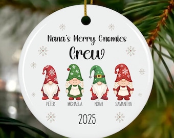 May include: A white ceramic Christmas ornament with the text "Nana's Merry Gnomies Crew" and the year 2025. It features four gnome illustrations with names below. The gnomes wear red, green, and white hats and outfits.