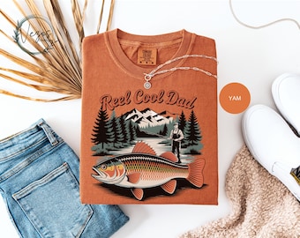 May include: An orange-colored t-shirt with the text "Reel Cool Dad" and a graphic of a fish, a fisherman, trees, and mountains. The shirt is folded and displayed with blue jeans, white shoes, and a necklace.