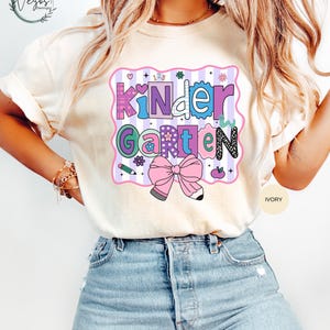 May include: Ivory-colored t-shirt with a colorful "Kindergarten" graphic. The graphic features a pink bow, flowers, and the words "Kindergarten" in various colors and fonts. The shirt is paired with blue jeans.