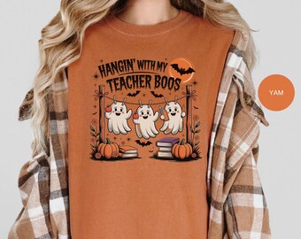 May include: Orange t-shirt with a Halloween-themed graphic. The design features three cartoon ghosts hanging on a clothesline with pumpkins, books, and bats. Text reads "Hangin' with my Teacher Boos."