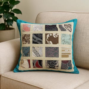 May include: Square decorative pillow with a patchwork design. The pillow features a teal border and a grid of fabric squares in various patterns and colors, including pink, black, and beige. The pillow is placed on a beige sofa.