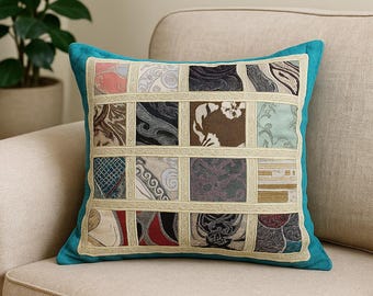May include: Square decorative pillow with a patchwork design. The pillow features a teal border and a grid of fabric squares in various patterns and colors, including pink, black, and beige. The pillow is placed on a beige sofa.