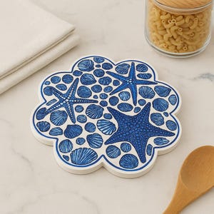 May include: A white, flower-shaped trivet with a blue seashell and starfish design. The trivet features a border and is decorated with various seashells and three blue starfish. A jar of pasta and a wooden spoon are in the background.