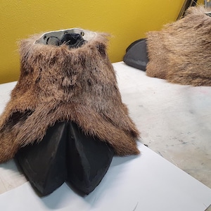 May include: A pair of brown faux fur animal feet with black hooves. The feet are designed to be worn as part of a costume. The fur is a mix of light and dark brown, and the hooves are a glossy black.