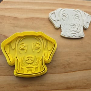 May include: A bright yellow plastic cookie cutter shaped like a dog head with floppy ears and a detailed snout, accompanied by a white embossed impression of the same dog face design, showing the facial features and texture of the canine.