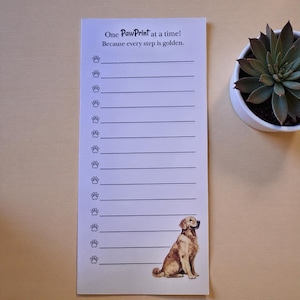 May include: A rectangular notepad with the text "One PawPrint at a time! Because every step is golden." features paw prints and a Golden Retriever illustration. The notepad has lined spaces for notes and is on a light-coloured surface.