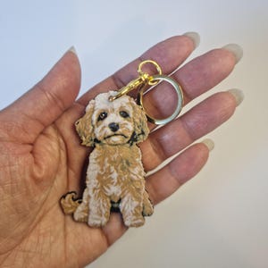 May include: A wooden dog keyring with a light brown and white coat, and a sad expression. The dog is seated and attached to a gold-coloured keyring and clasp. The keychain is held in a hand against a white background.