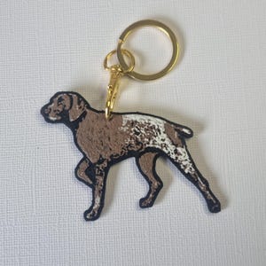 May include: A brown and white dog keyring with a gold-coloured ring and clasp. The dog is a German Shorthaired Pointer, with a black outline. A pet-themed accessory.