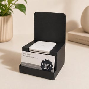 May include: A black business card holder with a white card reader and business cards. The business cards have the text "Cama 3D" and contact information. The holder is on a light-coloured surface.