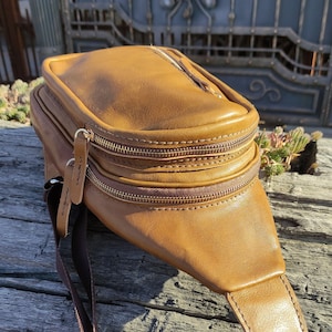 K&ouml;nnte beinhalten: Eine braune Leder-Bauchtasche mit einem braunen Gurt und goldenen Rei&szlig;verschl&uuml;ssen. Die Tasche hat zwei F&auml;cher und einen Klappenverschluss.