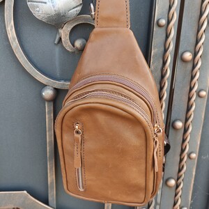 K&ouml;nnte beinhalten: Eine braune Leder-Umh&auml;ngetasche mit einem Rei&szlig;verschlussfach und einem braunen Trageriemen. Die Tasche h&auml;ngt an einem Metalltor.