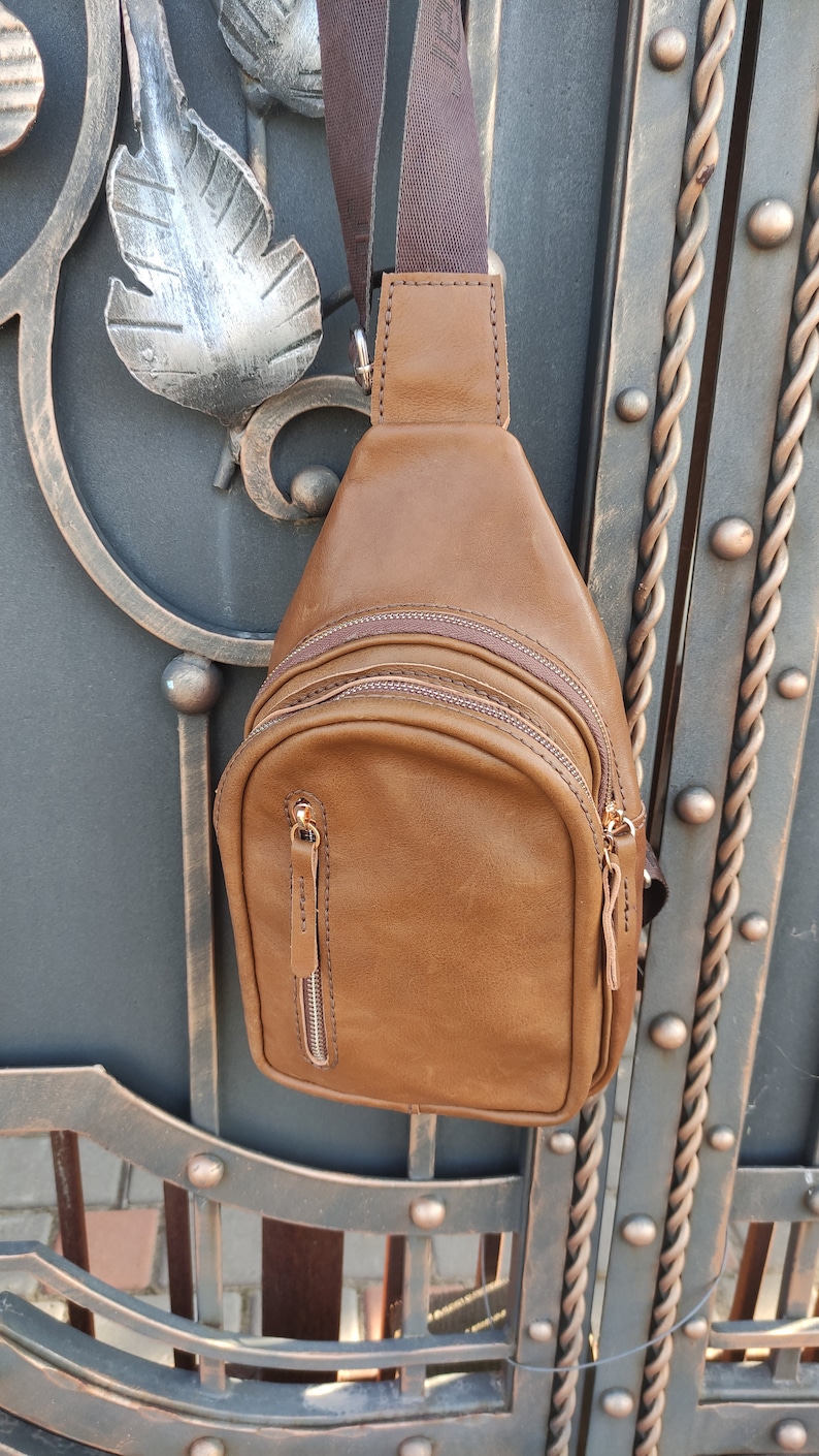 K&ouml;nnte beinhalten: Eine braune Leder-Umh&auml;ngetasche mit einem Rei&szlig;verschlussfach und einem braunen Trageriemen. Die Tasche h&auml;ngt an einem Metalltor.