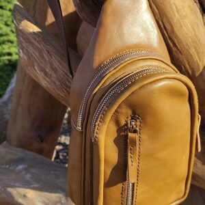 K&ouml;nnte beinhalten: Eine braune Leder-Umh&auml;ngetasche mit Rei&szlig;verschluss und einem langen, verstellbaren Trageriemen. Die Tasche h&auml;ngt an einer Holzoberfl&auml;che.