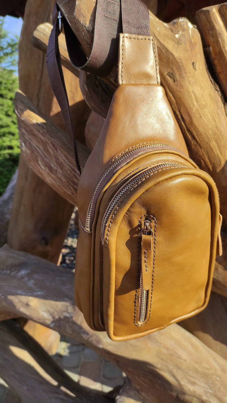 K&ouml;nnte beinhalten: Eine braune Leder-Umh&auml;ngetasche mit Rei&szlig;verschluss und einem langen, verstellbaren Trageriemen. Die Tasche h&auml;ngt an einer Holzoberfl&auml;che.