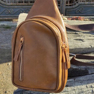 K&ouml;nnte beinhalten: Eine braune Leder-Umh&auml;ngetasche mit einer Rei&szlig;verschlusstasche und einem goldenen Rei&szlig;verschluss. Die Tasche liegt auf einer Holzoberfl&auml;che.
