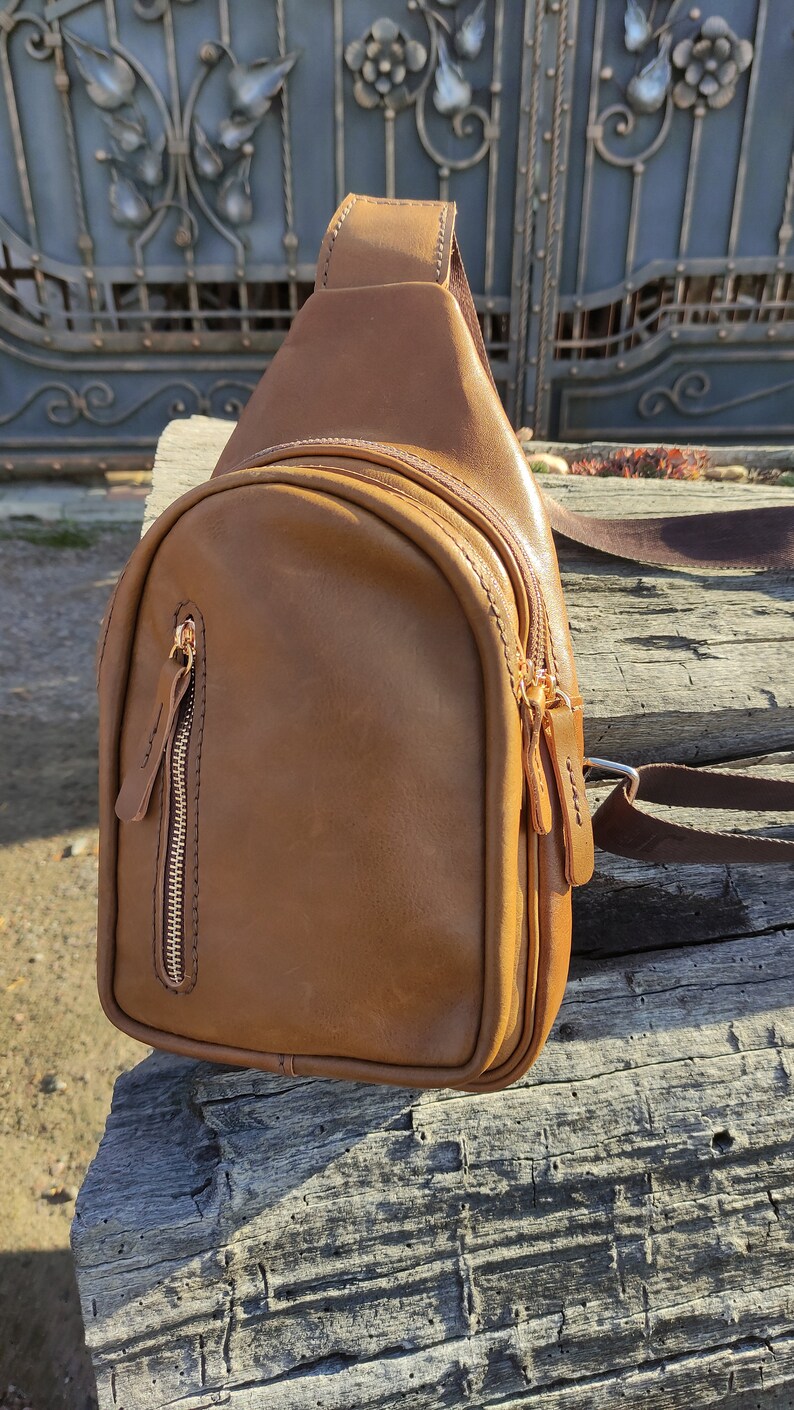 K&ouml;nnte beinhalten: Eine braune Leder-Umh&auml;ngetasche mit einer Rei&szlig;verschlusstasche und einem goldenen Rei&szlig;verschluss. Die Tasche liegt auf einer Holzoberfl&auml;che.
