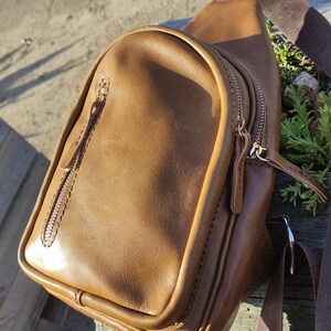 K&ouml;nnte beinhalten: Eine braune Leder-Umh&auml;ngetasche mit zwei Rei&szlig;verschlussf&auml;chern und einem verstellbaren braunen Trageriemen. Die Tasche liegt auf einer Holzoberfl&auml;che.