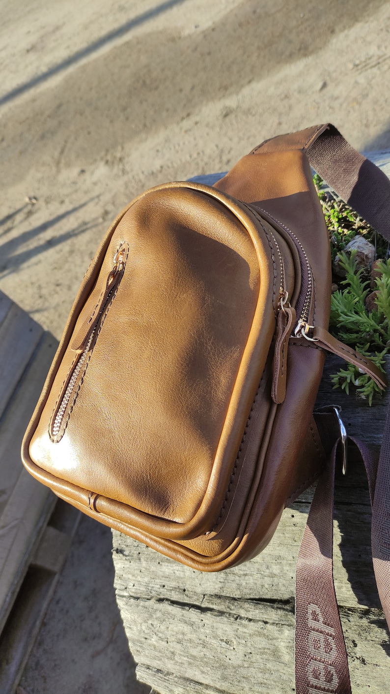 K&ouml;nnte beinhalten: Eine braune Leder-Umh&auml;ngetasche mit zwei Rei&szlig;verschlussf&auml;chern und einem verstellbaren braunen Trageriemen. Die Tasche liegt auf einer Holzoberfl&auml;che.
