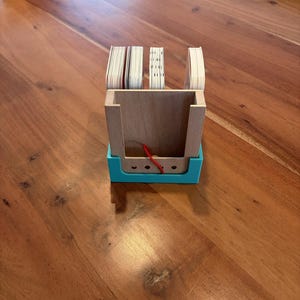 Könnte beinhalten: Ein Visitenkartenhalter aus Holz mit türkisfarbenem Sockel. Der Halter enthält mehrere Kartenstapel. Am Boden des Halters ist eine rote Schnur zu sehen. Der Halter steht auf einem Holztisch.