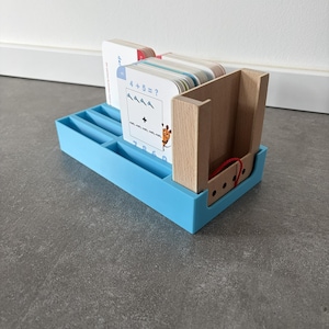 Könnte beinhalten: Ein blauer Kunststoff-Organizer mit Fächern enthält Kartenhalter aus Holz und einen Stapel Mathe-Lernkarten. Die Lernkarten zeigen Additionsaufgaben und eine Giraffenillustration. Der Kartenhalter aus Holz hat ein rotes Gummiband.