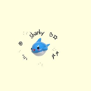 May include: A blue and white shark figurine with the word "sharky" written above it. The shark has a cute, cartoonish design with a red blush and black eyes. Surrounding the shark are doodle-like stars and hearts.