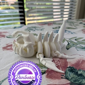 May include: White 3D printed skeletal creature with wings, lying on a floral fabric. The creature has a large head with teeth and a long tail. The image includes a purple and white logo with the text "Authorized Seller Printverse Authorized Seller".