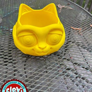 May include: A bright yellow cat-shaped planter with large, round eyes and a smiling face. The planter has a wide opening for plants and is made of a solid material. The Flexi Factory authorized seller logo is visible.