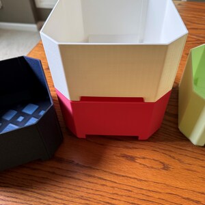 May include: A collection of geometric planters in various colors. The largest planter is white with a red base. A black planter and a green planter are also visible. The planters are made of a solid material and have a modern design.