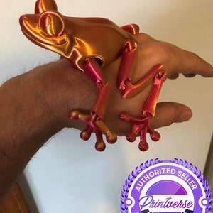 May include: A 3D printed articulated frog figurine with a metallic gold and pink finish. The frog is posed with its legs outstretched and its head tilted slightly to the side. The figurine is sitting on a person's hand.