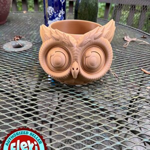 May include: A brown owl-shaped planter with large, detailed eyes and feather-like accents. The planter is made of a light brown material and sits on a metal table. The Flexi Factory logo is visible in the bottom left corner.