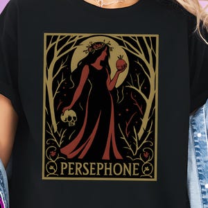 May include: Black t-shirt featuring a gold and red Persephone graphic. The design includes a woman holding a skull and an apple, with the name "PERSEPHONE" below. The shirt has a crew neck and short sleeves.