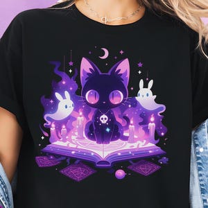 Puede incluir: Camiseta negra con un gato de dibujos animados de ojos grandes, sentado en un libro abierto con velas, fantasmas y una luna creciente. El gato tiene un collar de calavera. La palabra "BLACK" está en un círculo en la parte inferior.
