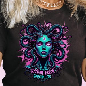 May include: Black t-shirt with a striking graphic of a Medusa-inspired design. The image features a teal-skinned woman with snake hair and pink highlights, along with the text "SYSTEM ERROR GORGON.EXE". The shirt has short sleeves.