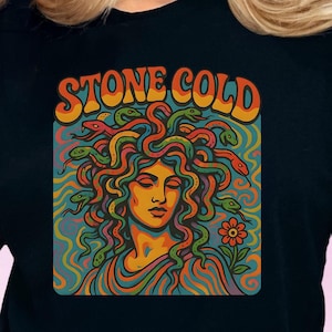 Könnte beinhalten: Schwarzes T-Shirt mit einem farbenfrohen Grafikdesign von Medusa mit Schlangen, einer Blume und dem Text "STONE COLD" in Orange und Gelb. Das Design hat einen Retro-Psychedelic-Vibe.