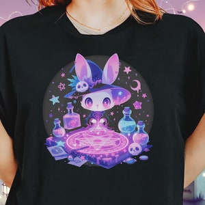 May include: A black t-shirt with a graphic of a white rabbit wearing a witch's hat and holding a wand. The rabbit is surrounded by purple and pink potion bottles and a pentagram.