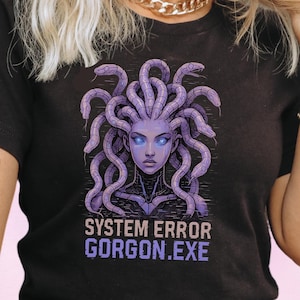 Puede incluir: Camiseta negra con un diseño de Medusa, representada con serpientes moradas como cabello y ojos azules. El texto "SYSTEM ERROR GORGON.EXE" está impreso debajo de la ilustración. La camiseta es de cuello redondo y manga corta.