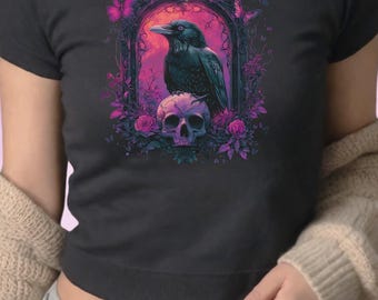 May include: Black t-shirt featuring a gothic design. The graphic includes a raven perched above a skull, framed by a floral arch with butterflies. The artwork uses shades of black, purple, and pink.