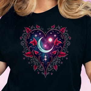 Op de afbeelding: Zwart T-shirt met een hartvormig ontwerp met een maan, sterren en een nevel. Het ontwerp is omlijst door rode bladeren en decoratieve krullen, wat een gotische en mystieke esthetiek creëert.
