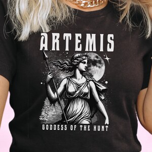 May include: Black t-shirt featuring a detailed illustration of Artemis, the Greek goddess of the hunt, holding a spear. The design includes the word "ARTEMIS" above the image and "GODDESS OF THE HUNT" below.