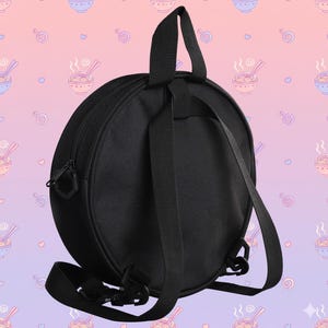 May include: Black round backpack with adjustable shoulder straps and a top handle. The backpack has a zippered closure and a solid black design. The background features a repeating pattern of ramen bowls and hearts.