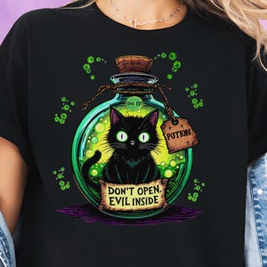 May include: Black t-shirt with a cartoon image of a black cat in a green potion bottle. The bottle has a cork stopper and a wooden tag that says "Potion." The shirt also has the text "Don't Open, Evil Inside."