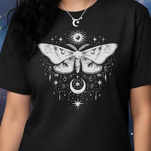 Op de afbeelding: Zwart t-shirt met een gedetailleerde witte mot illustratie met hemelse elementen zoals sterren, een zon en een maan. Het ontwerp is gecentreerd op het shirt. Een zilveren ketting met een maan hanger is ook zichtbaar.