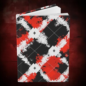 K&ouml;nnte beinhalten: Hardcover-Journal mit einem auff&auml;lligen Argyle-Muster in Schwarz, Rot und Wei&szlig;. Das Design zeigt ein Diamantgitter mit Farbspritzern und kleinen Diamantformen. Das Journal ist ge&ouml;ffnet und zeigt wei&szlig;e Seiten.