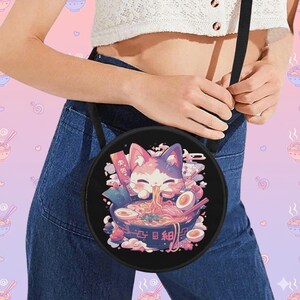 May include: Black round crossbody bag featuring a cartoon cat enjoying ramen. The cat is pink and white, with a bowl of ramen in front of it. The bag has a black strap and is worn over the shoulder.