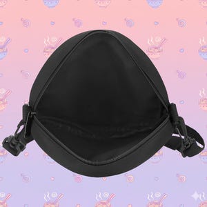 May include: Black messenger bag with an open zipper, revealing the interior. The bag has a rectangular shape with rounded corners and a black adjustable strap. The background features a repeating pattern of ramen bowls and hearts.