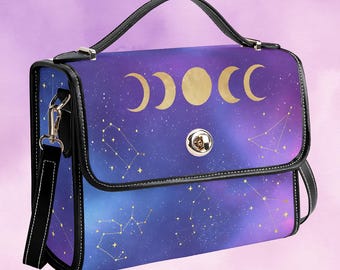 May include: A satchel-style handbag with a celestial design. The bag features a handle, a detachable shoulder strap, and a clasp closure. The front panel displays a purple and blue galaxy pattern with gold crescent moons, a full moon, and constellations.