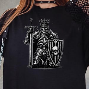 Può includere: T-shirt nero con grafica in bianco e nero di un cavaliere scheletrico in armatura, con spada e scudo. Il cavaliere indossa una corona e lo scudo mostra un teschio. Design gotico e dettagliato.