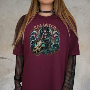 May include: Burgundy t-shirt featuring a detailed graphic of a mermaid seated on a skull, with the text "SEA WITCH" above. The mermaid has long dark hair and a green tail, surrounded by stylized waves and glowing blue accents.
