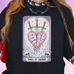 Pode incluir: Camiseta preta de manga comprida com um design de carta de tarô. A carta mostra um coração rosa rachado perfurado por três espadas, com o texto "THREE OF SWORDS". O design inclui caveiras e detalhes florais.