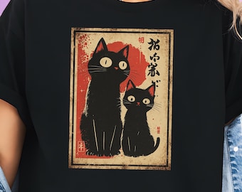Op de afbeelding: Zwart T-shirt met een retro grafische afbeelding van twee zwarte katten met grote ogen, tegen een rode zonsondergang achtergrond. Japanse tekst is zichtbaar boven de katten. Het ontwerp heeft een vintage, artistieke uitstraling.